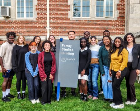 Cohort of 14 first year graduate students pose next to Family Studies Building Sign